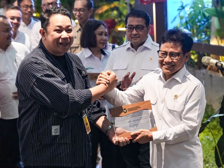 Two men shake hands while presenting a glass award plaque; the recipient in a white uniform smiles as onlookers watch in the background.