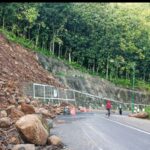 Road blocked by a landslide with large rocks scattered across the lane a few people and a motorcycle nearby on a curving mountain road