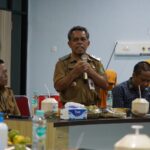 A man in a tan official uniform speaks into a microphone at a conference table surrounded by colleagues in similar attire and yellow headscarves with food and beverages on the table