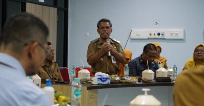 A man in a tan official uniform speaks into a microphone at a conference table surrounded by colleagues in similar attire and yellow headscarves with food and beverages on the table