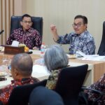 People in a formal meeting around a conference table two men in batik shirts are at the head one gesturing as he speaks
