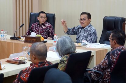 People in a formal meeting around a conference table two men in batik shirts are at the head one gesturing as he speaks