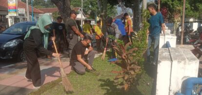 Community volunteers clean a street garden area with brooms and tools, gathered near plants and a parked car in the background.