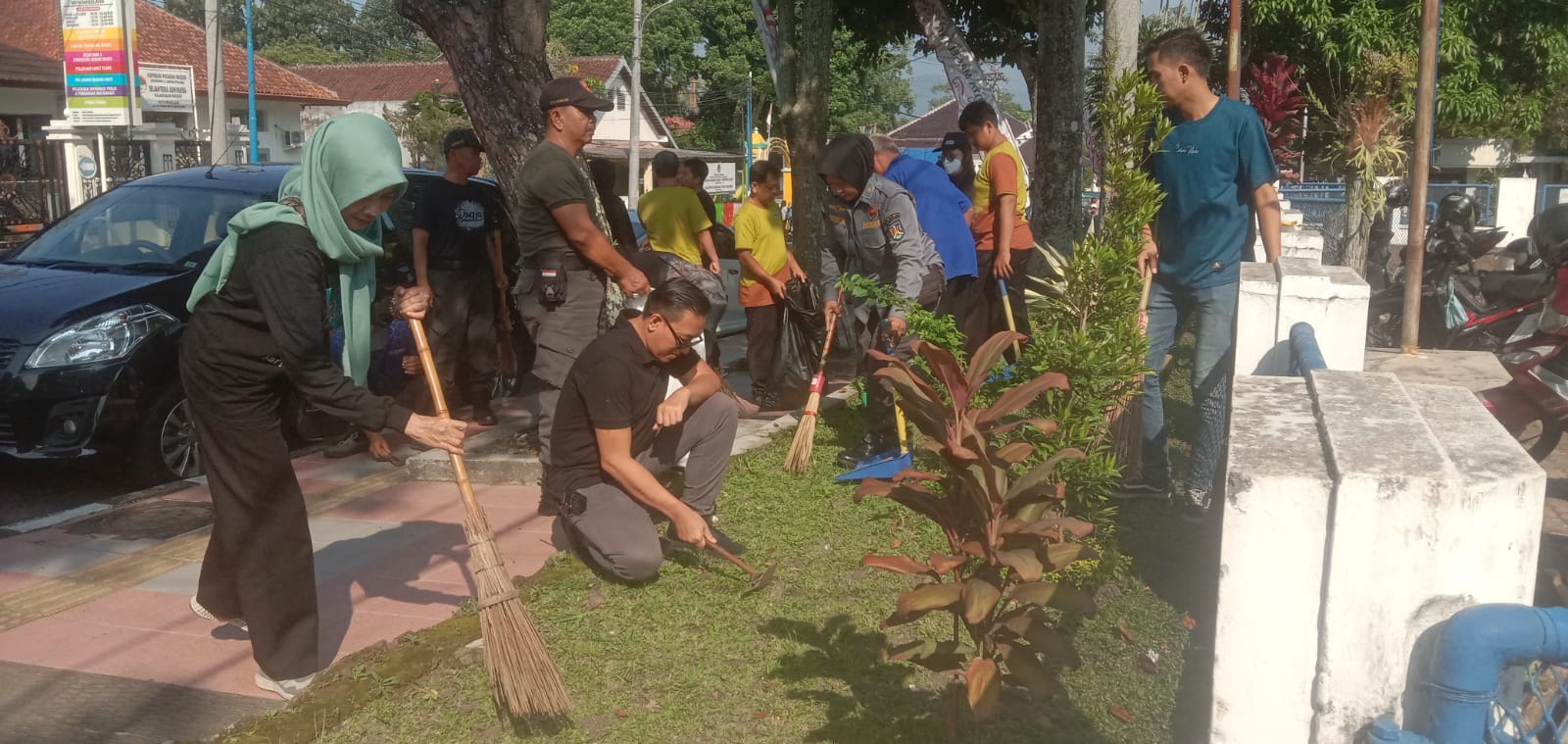Community volunteers clean a street garden area with brooms and tools gathered near plants and a parked car in the background