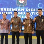 Four men in batik shirts pose with fists raised on a stage during a ceremony in front of a blue banner reading peresmian digital election lab logo backdrop