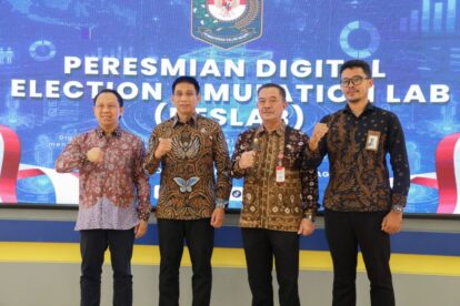 Four men in batik shirts pose with fists raised on a stage during a ceremony in front of a blue banner reading 'Peresmian Digital Election Lab' (logo backdrop).