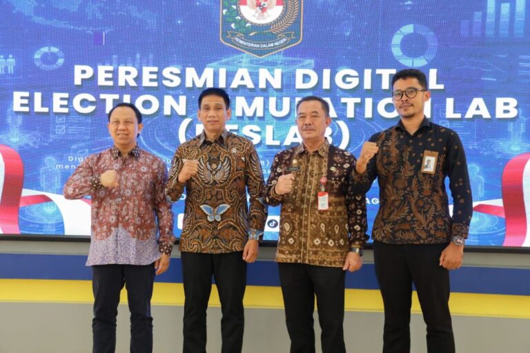 Four men in batik shirts pose with fists raised on a stage during a ceremony in front of a blue banner reading 'Peresmian Digital Election Lab' (logo backdrop).