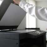 Person in a white shirt lifts the lid of a large photocopier while reaching the control panel in an office setting