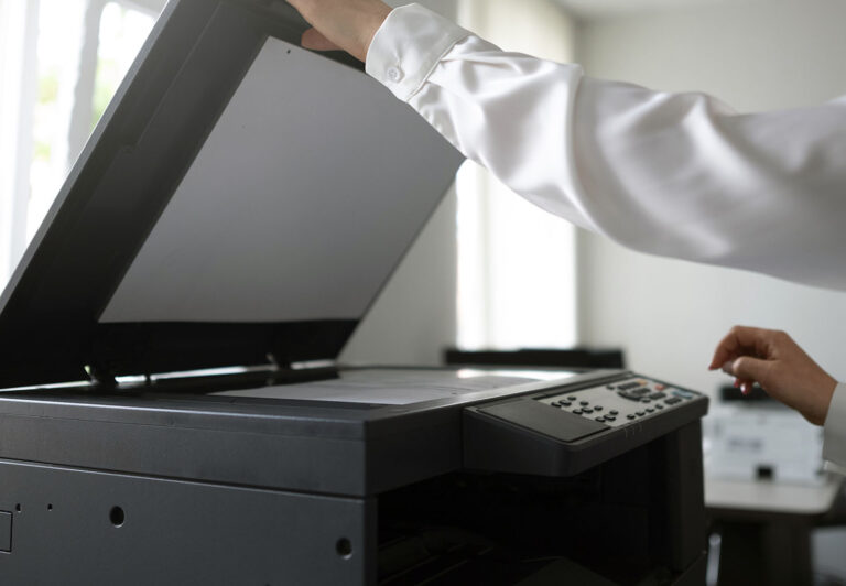 Person in a white shirt lifts the lid of a large photocopier while reaching the control panel in an office setting.