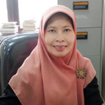 Smiling woman wearing a pink hijab and a jeweled brooch seated at a desk with a filing cabinet and stacks of papers in the background