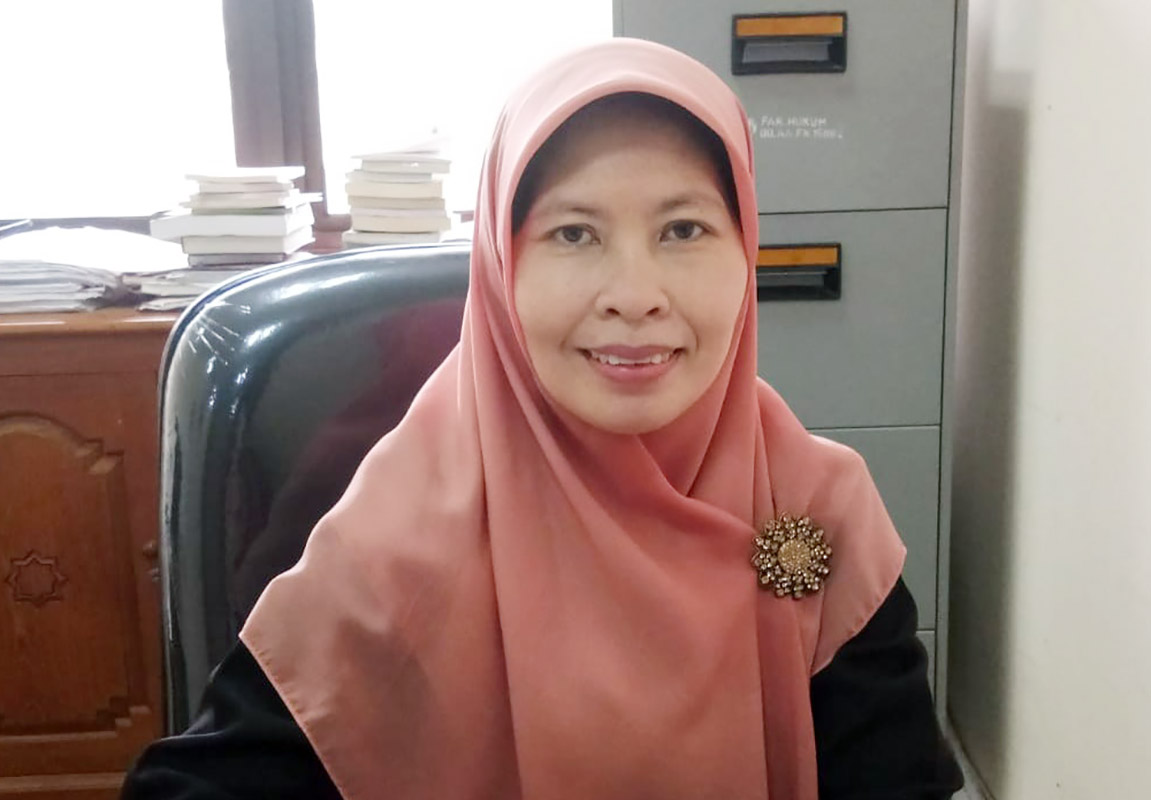 Smiling woman wearing a pink hijab and a jeweled brooch seated at a desk with a filing cabinet and stacks of papers in the background