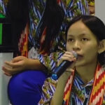 Young woman with short black hair speaks into a microphone at a conference wearing a colorful patterned shirt and red lanyard