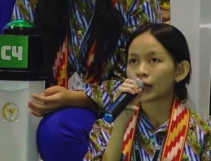 Young woman with short black hair speaks into a microphone at a conference wearing a colorful patterned shirt and red lanyard