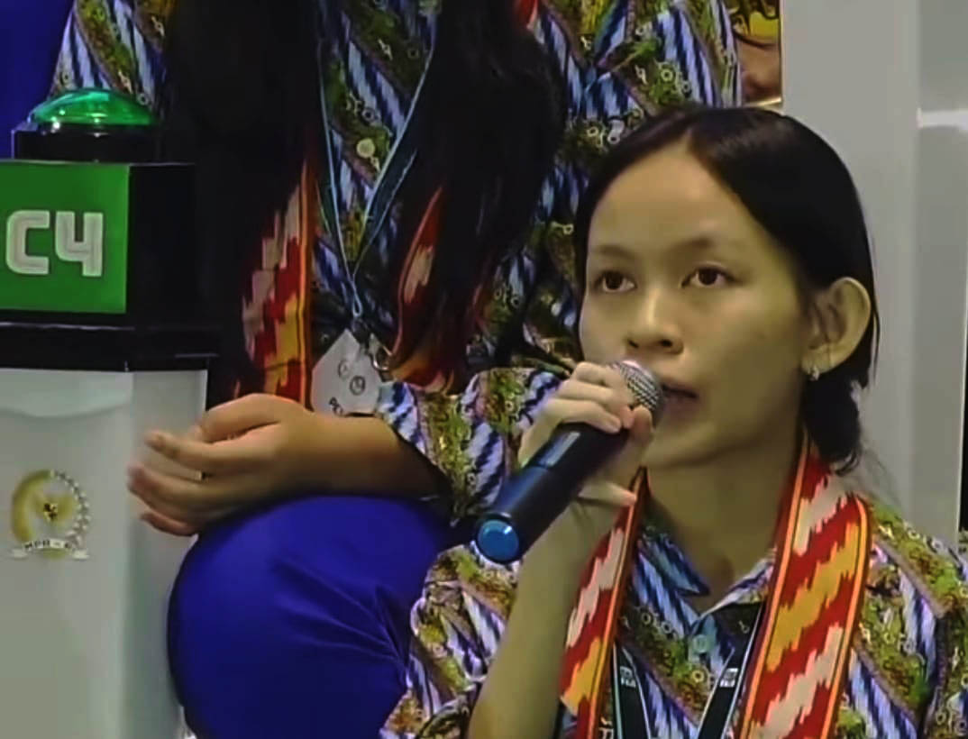 12ejosepha catrawarta Young woman with short black hair speaks into a microphone at a conference wearing a colorful patterned shirt and red lanyard