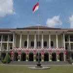 Front view of a neoclassical campus building with tall columns arches and a central flagpole flying a red and white flag with decorative banners along the facade