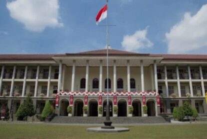 Front view of a neoclassical campus building with tall columns, arches, and a central flagpole flying a red-and-white flag, with decorative banners along the facade.