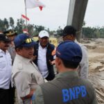 Group of indonesian officials in uniform and caps inspecting a constructionsite with sand piles and a damaged riverbank flags in the background