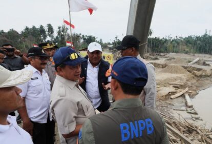 Group of indonesian officials in uniform and caps inspecting a constructionsite with sand piles and a damaged riverbank flags in the background