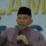Older man wearing a black cap speaks into a handheld microphone while gesturing with his left hand on stage