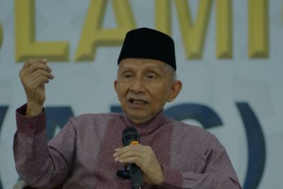 Older man wearing a black cap speaks into a handheld microphone while gesturing with his left hand on stage.