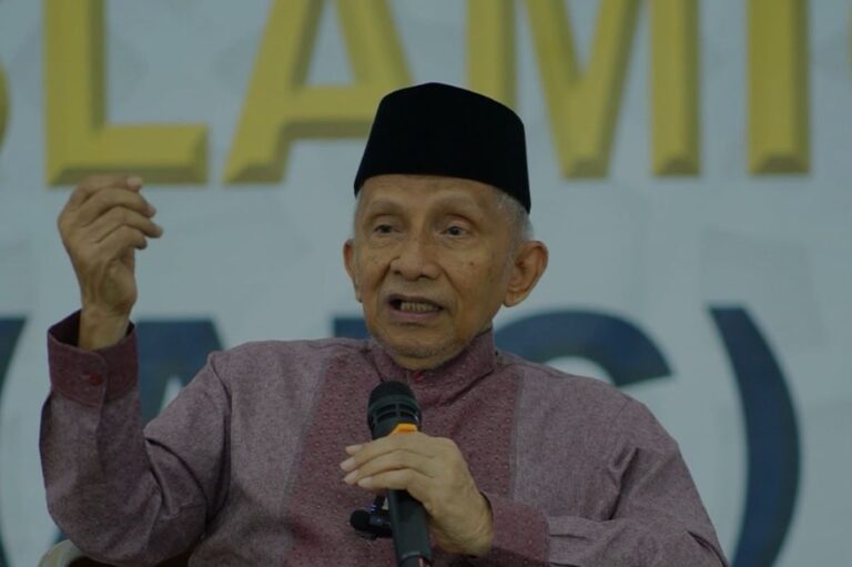 Older man wearing a black cap speaks into a handheld microphone while gesturing with his left hand on stage.