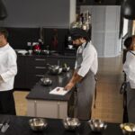Three chefs work in a modern kitchen one opening a tall fridge another chopping meat at a counter and a third reaching for a pot overhead