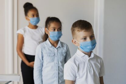 Three children in a hallway wearing blue face masks, lined up in a row.