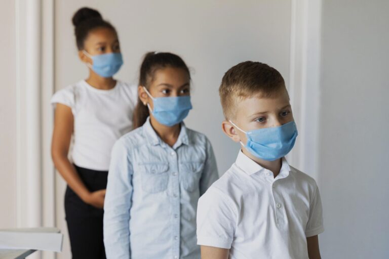 Three children in a hallway wearing blue face masks, lined up in a row.