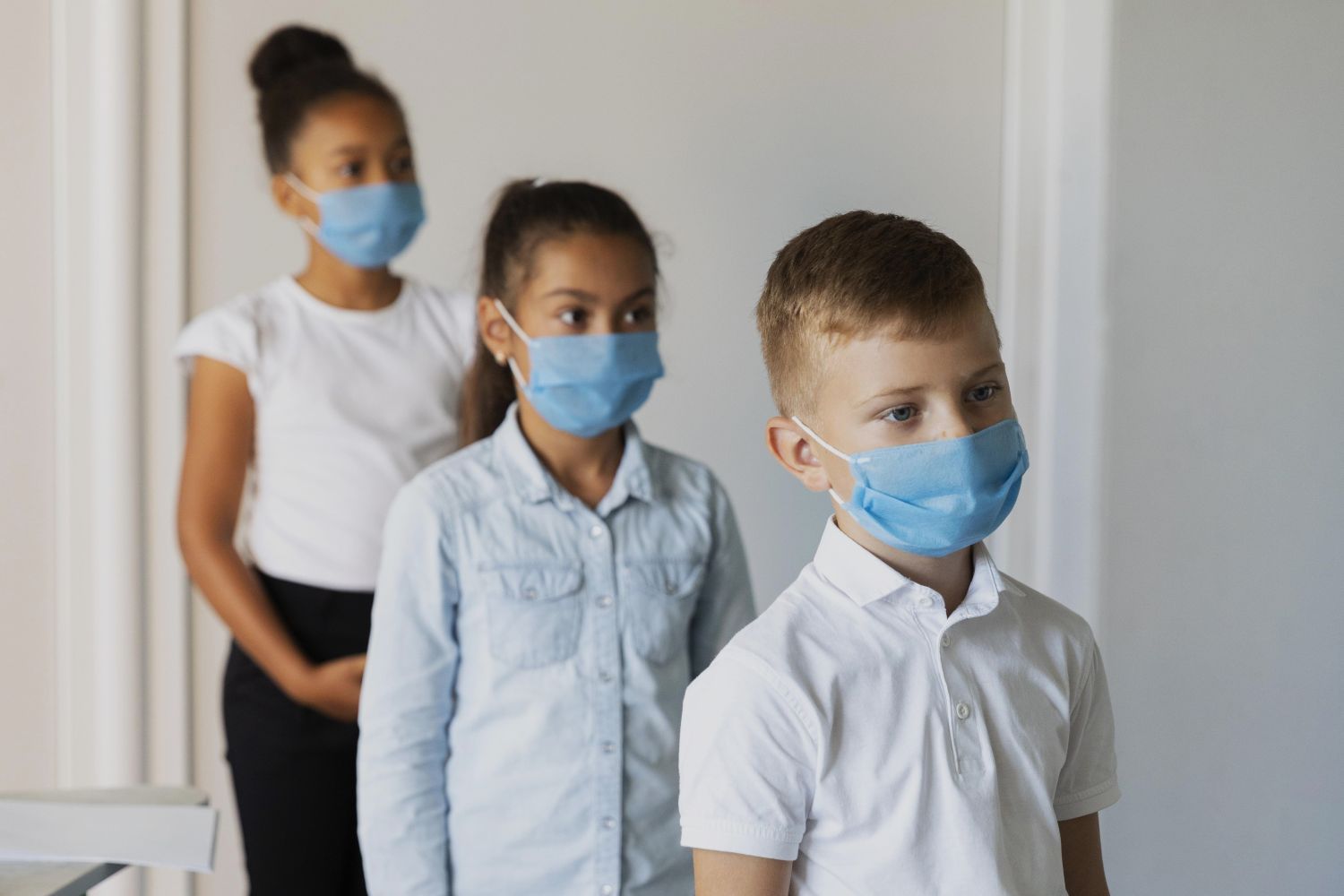 Three children in a hallway wearing blue face masks lined up in a row