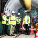 Group of construction workers in hard hats and high visibility vests standing inside a tunnel with a large yellow boring machine part overhead posing for a photo inside a work zone