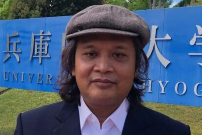 Man in a flat cap and dark blazer posing in front of a bright blue university sign with large characters, outdoor setting.