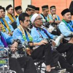 Group of men in blue traditional outfits and black headwear seated in chairs and wheelchairs at an event some wearing scarves with a security official in a beige vest in the background