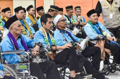 Group of men in blue traditional outfits and black headwear seated in chairs and wheelchairs at an event some wearing scarves with a security official in a beige vest in the background