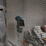 Medical worker in full protective ppe and face shield stands outside a patient room observing a patient in bed behind horizontal blinds