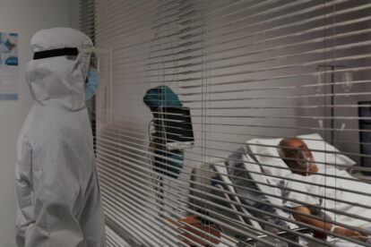 Medical worker in full protective PPE and face shield stands outside a patient room, observing a patient in bed behind horizontal blinds.