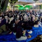Large crowd of people seated on blue tarps under tents at night many wearing hijabs attending an outdoor gathering with green stage lights