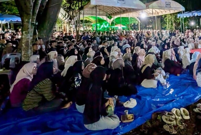 Large crowd of people seated on blue tarps under tents at night, many wearing hijabs, attending an outdoor gathering with green stage lights.
