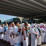 Group of muslim pilgrims in white ihram robes wearing green scarves line up beneath a highway overpass with green buses in the background