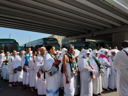 Group of muslim pilgrims in white ihram robes wearing green scarves line up beneath a highway overpass with green buses in the background