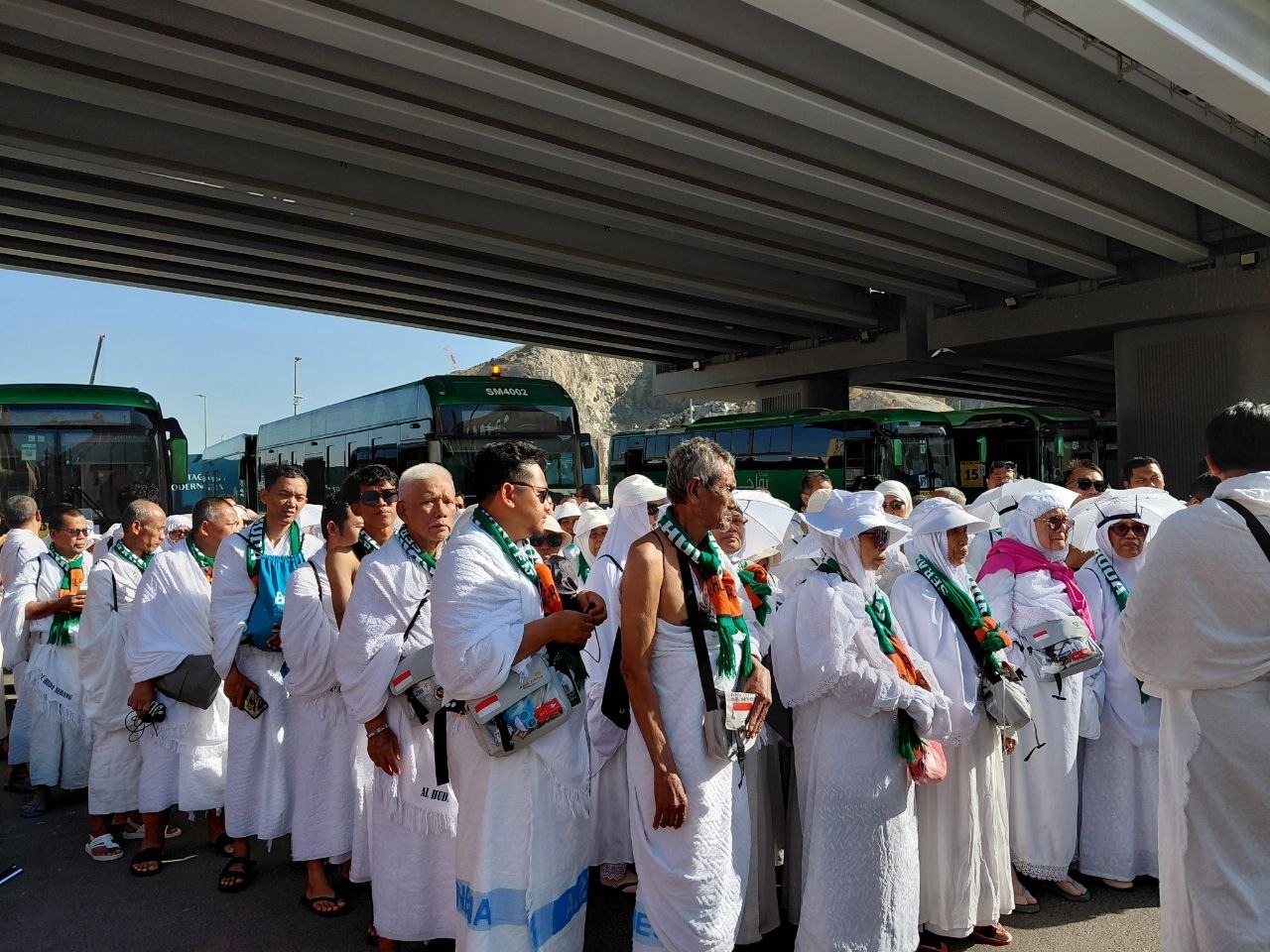 Group of muslim pilgrims in white ihram robes wearing green scarves line up beneath a highway overpass with green buses in the background