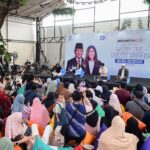 Stage panel discussion at a public event with five speakers seated on cushions a large banner behind features indonesian text and images of a man and woman audience sits on the floor and on beanbags in a bright plant filled venue