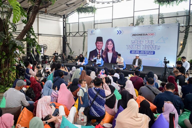 Stage panel discussion at a public event with five speakers seated on cushions; a large banner behind features Indonesian text and images of a man and woman; audience sits on the floor and on beanbags in a bright, plant-filled venue.