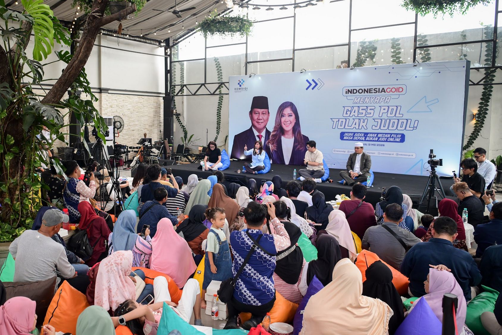 Stage panel discussion at a public event with five speakers seated on cushions a large banner behind features indonesian text and images of a man and woman audience sits on the floor and on beanbags in a bright plant filled venue