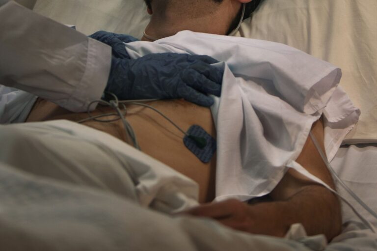 Medical professional wearing blue gloves resting a hand on a patient’s back in a hospital bed
