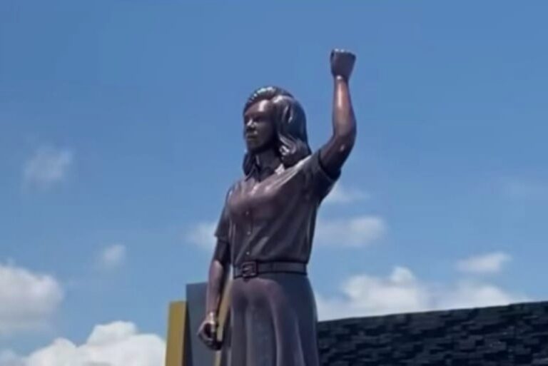 Bronze statue of a man with his right fist raised, outdoors against a blue sky with clouds.