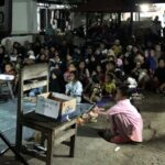 Large night time gathering of families seated on the ground for a charity event with a cardboard box labeled anak untuk papua in the foreground