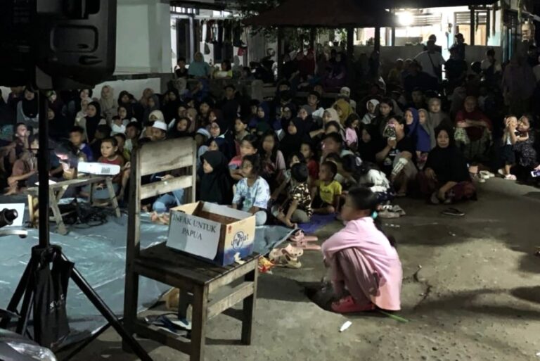 Large night-time gathering of families seated on the ground for a charity event, with a cardboard box labeled 'ANAK UNTUK PAPUA' in the foreground.