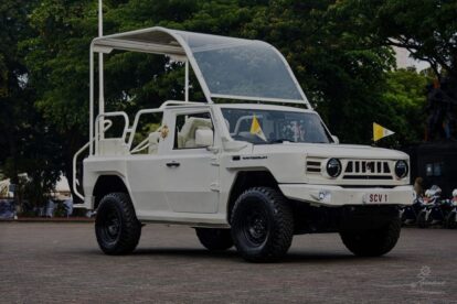 White vintage off-road SUV with a clear curved canopy, parked in an open square during an event.