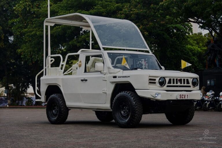 White vintage off-road SUV with a clear curved canopy, parked in an open square during an event.