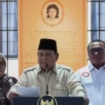 Three people at a podium during a press conference center man speaks into a microphone flanked by two colleagues against an orange wall with a portrait